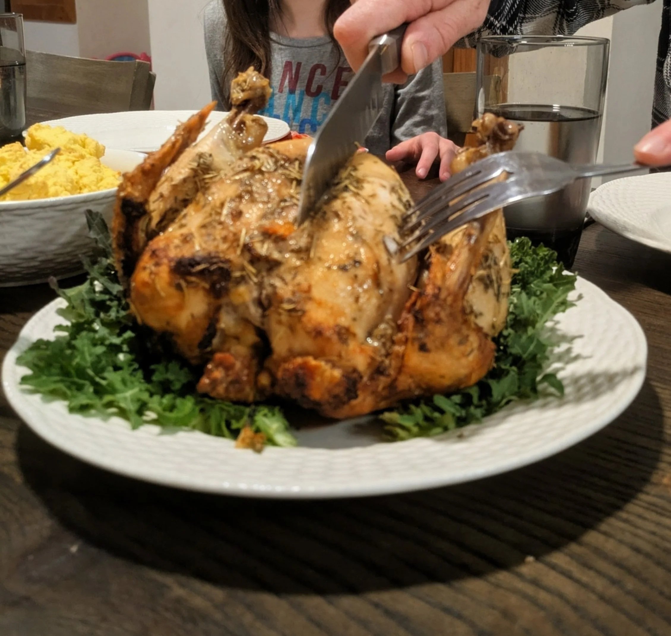 A perfectly roasted chicken being carved on a plate garnished with fresh greens, with a child visible in the background, showcasing a family meal setting.