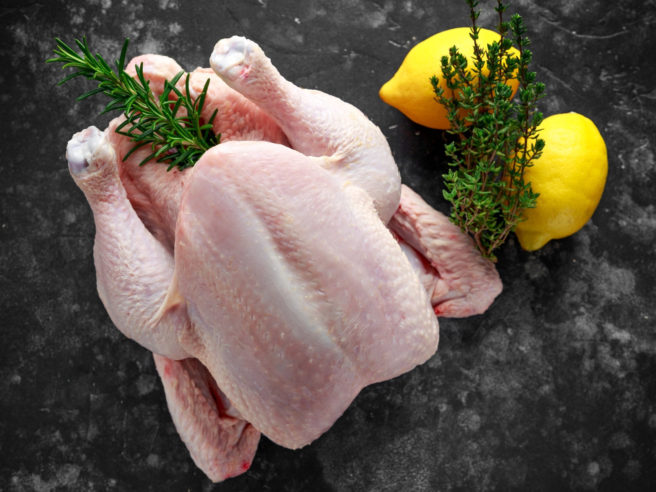 chicken_raw_whole_lemon_thyme_ (2) Raw whole chicken with fresh rosemary and thyme, accompanied by two lemons, on a dark textured surface, ideal for cooking or recipe preparation.