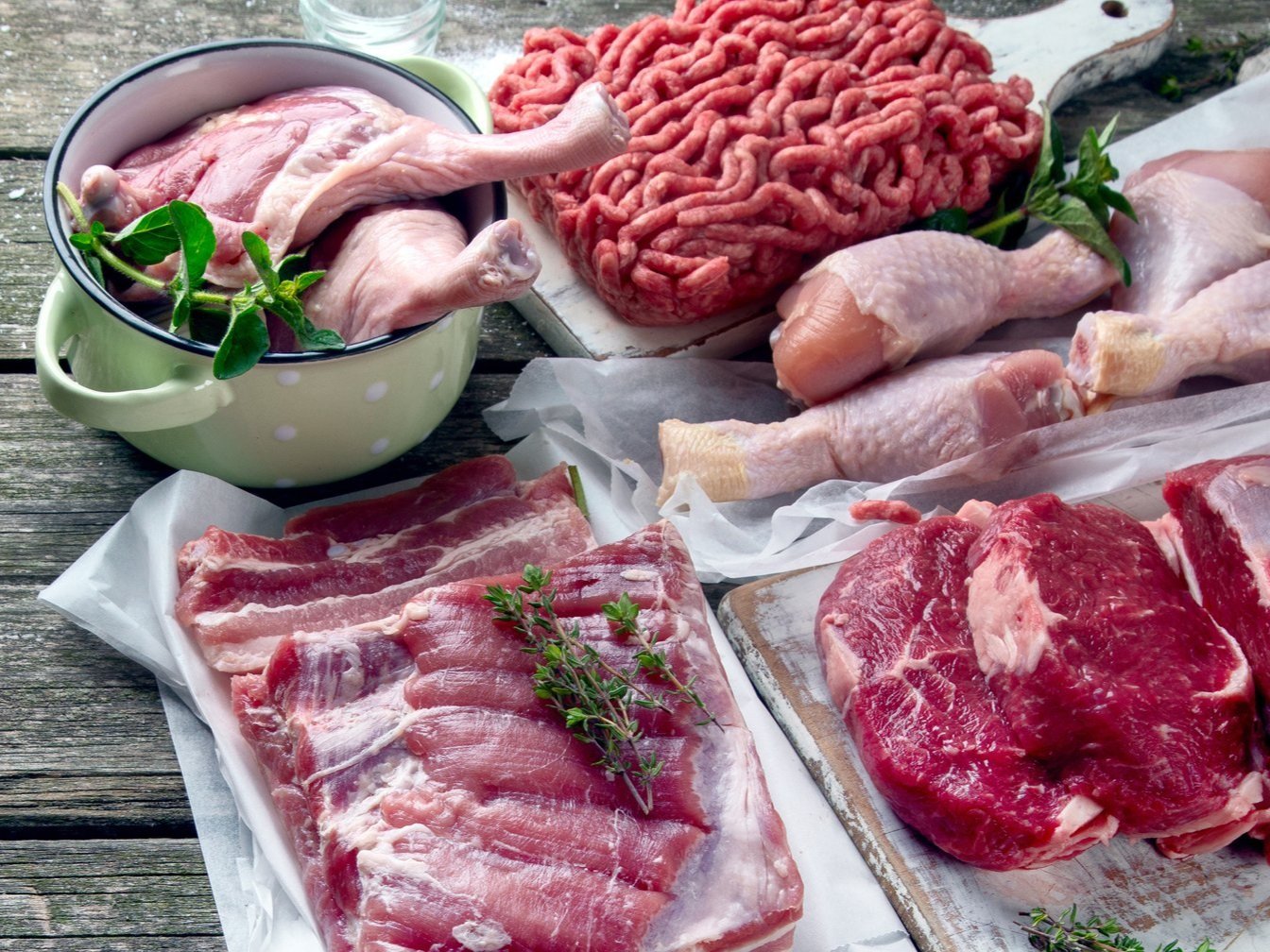 meat_raw_variety_beef_pork_poultry Assorted fresh meats displayed on a wooden surface, including chicken legs in a green bowl, ground beef, pork belly slices, and beef cuts garnished with fresh herbs.