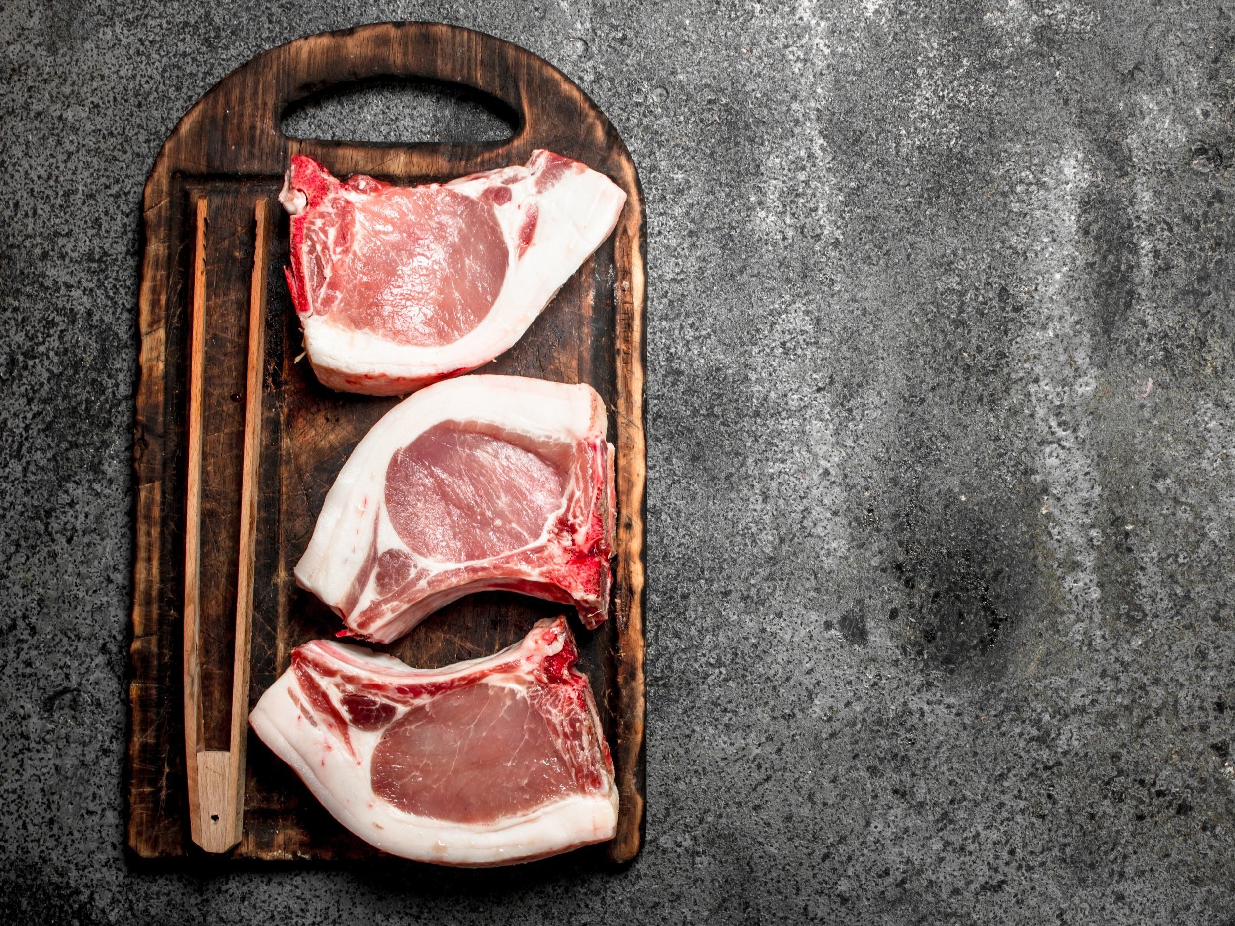 pork_chops_raw_cutting_board (2) Pork chops displayed on a wooden cutting board with cooking tongs, set against a textured gray background.