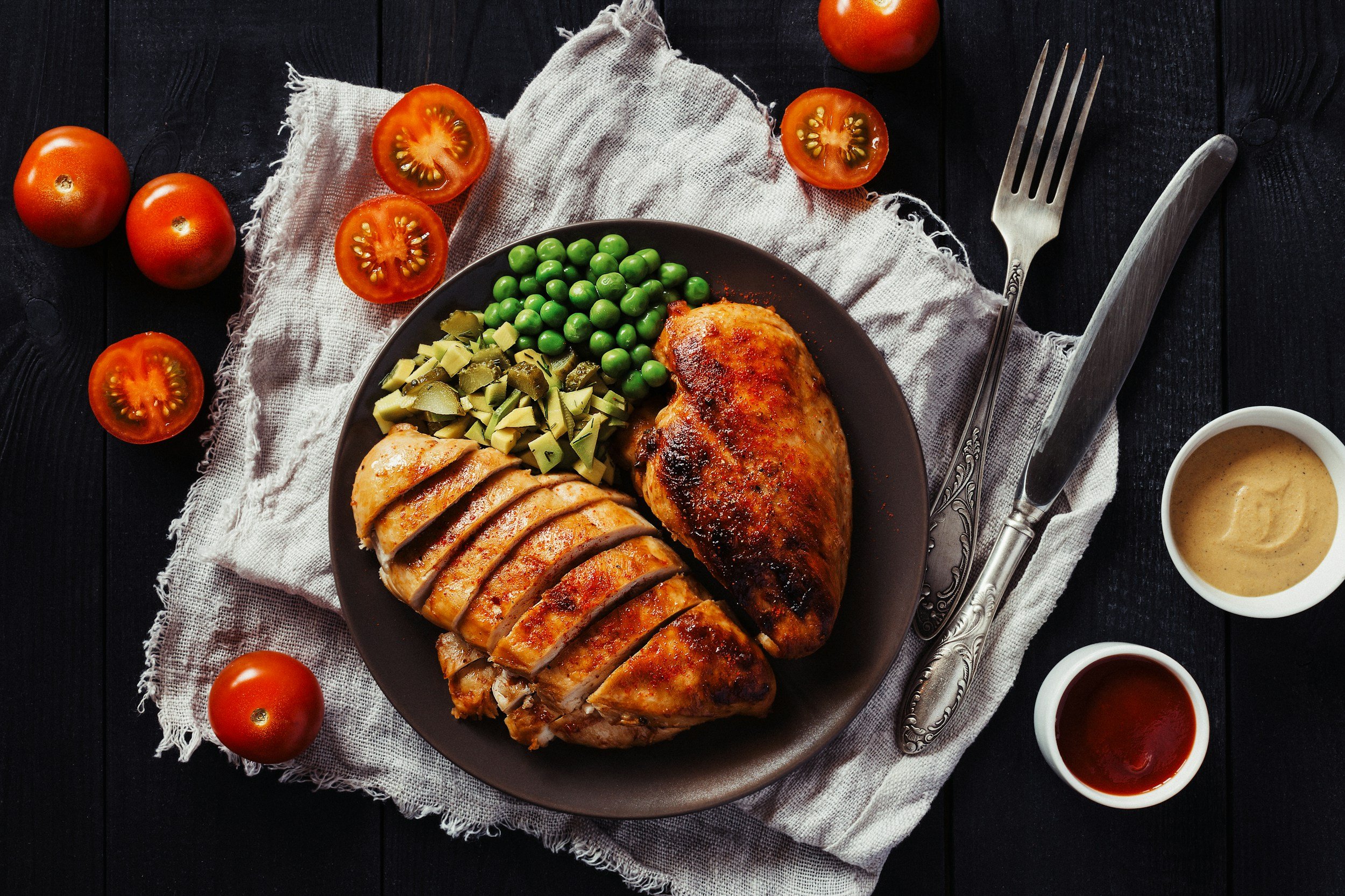 unsplash-image-DBVIb_rWUKw Grilled chicken breast served with green peas, diced artichokes, and fresh tomatoes on a rustic linen napkin, accompanied by two dipping sauces in small bowls.