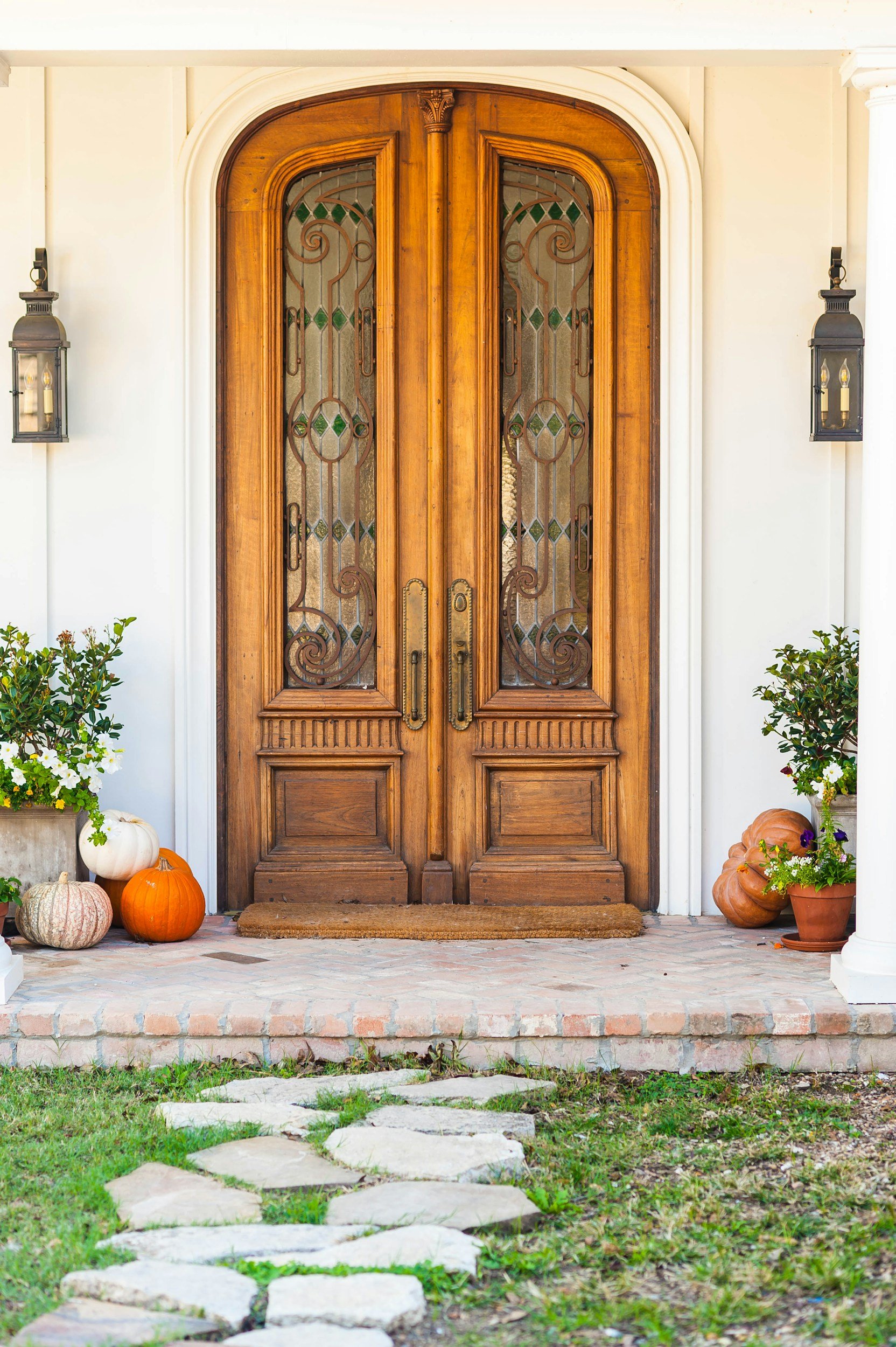 unsplash-image-LzOTAqHlNw8 Elegant wooden double doors with stained glass accents, flanked by lanterns, lead to a charming entryway adorned with seasonal pumpkins and vibrant flowers in pots. A stone pathway guides the way to the welcoming porch.