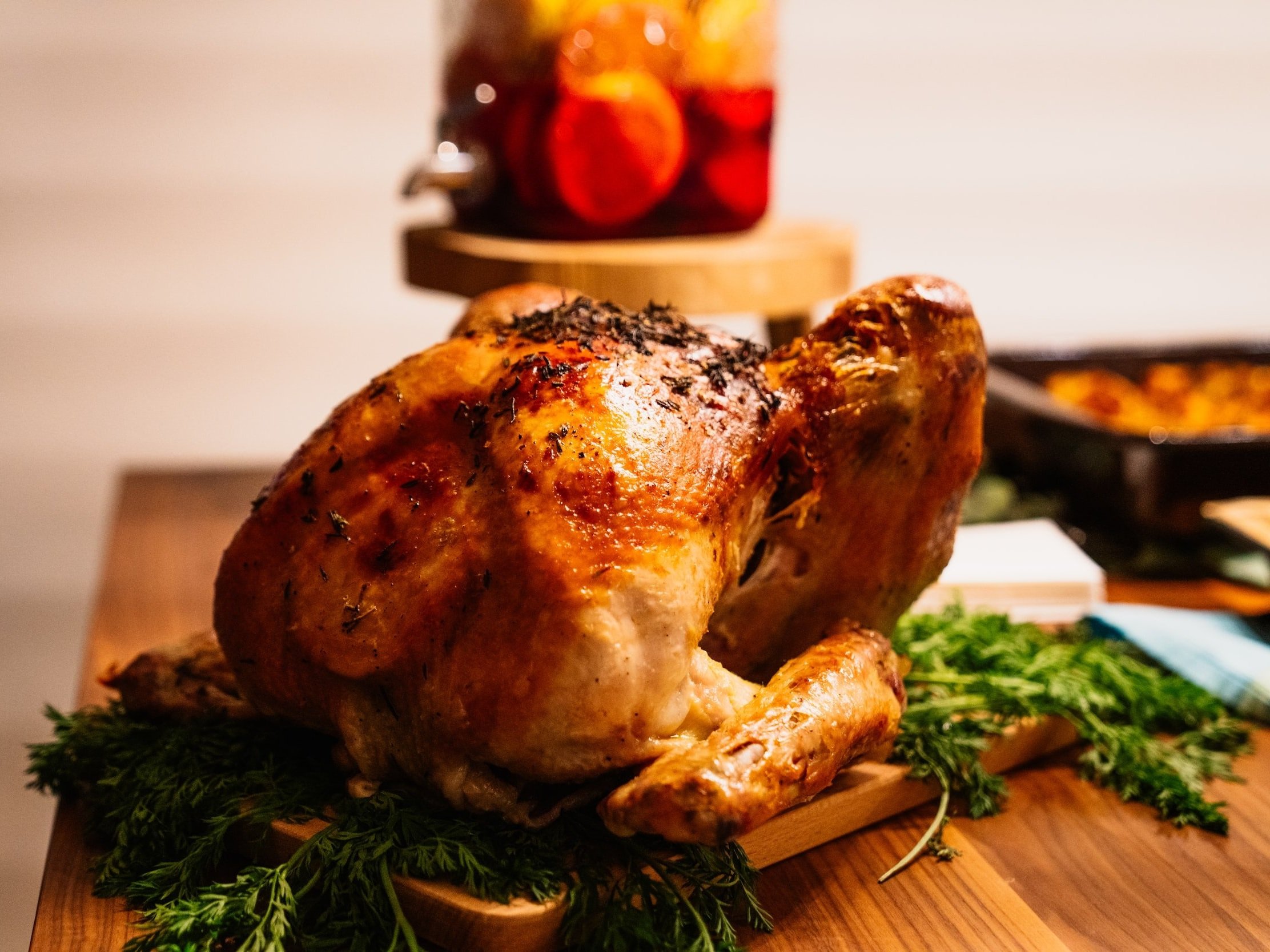 Roasted turkey garnished with fresh herbs, displayed on a wooden cutting board, with a colorful jar of fruit-infused drink in the background, perfect for holiday feasts or family gatherings.